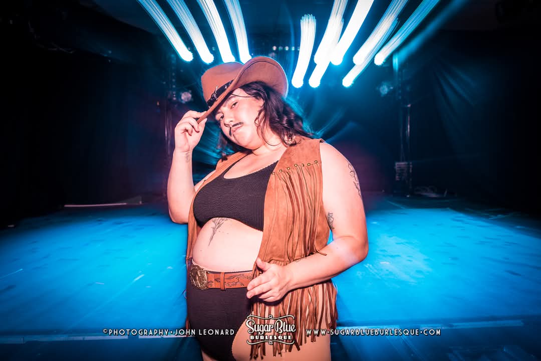 Ash on stage in a playful cowboy-inspired burlesque look, including a drawn on moustache. With one hand on the hat brim and the other on their hip, they strike a confident, campy pose.